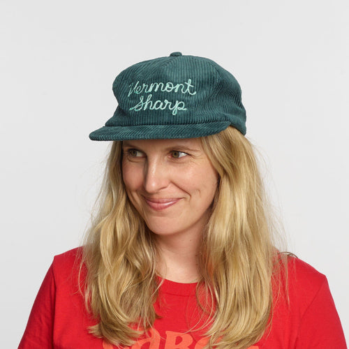 A woman with long blonde hair smiles against a white background, wearing a teal Vermont Sharp Chainstitched Corduroy Hat by New Duds and a red shirt.