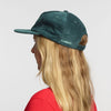 A person with long blonde hair wears the New Duds Vermont Sharp Chainstitched Corduroy Hat—a dark green five-panel cap with “CABOT” chain-stitched on the side—plus a red shirt, viewed from the side and back against a plain light background.