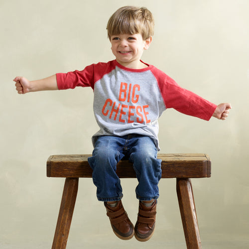 A young boy sits smiling on a bench in a gray and red Big Cheese Toddler T-Shirt by New Duds, extending his arms. The soft cotton fabric pairs perfectly with his blue jeans and brown shoes against a plain backdrop.