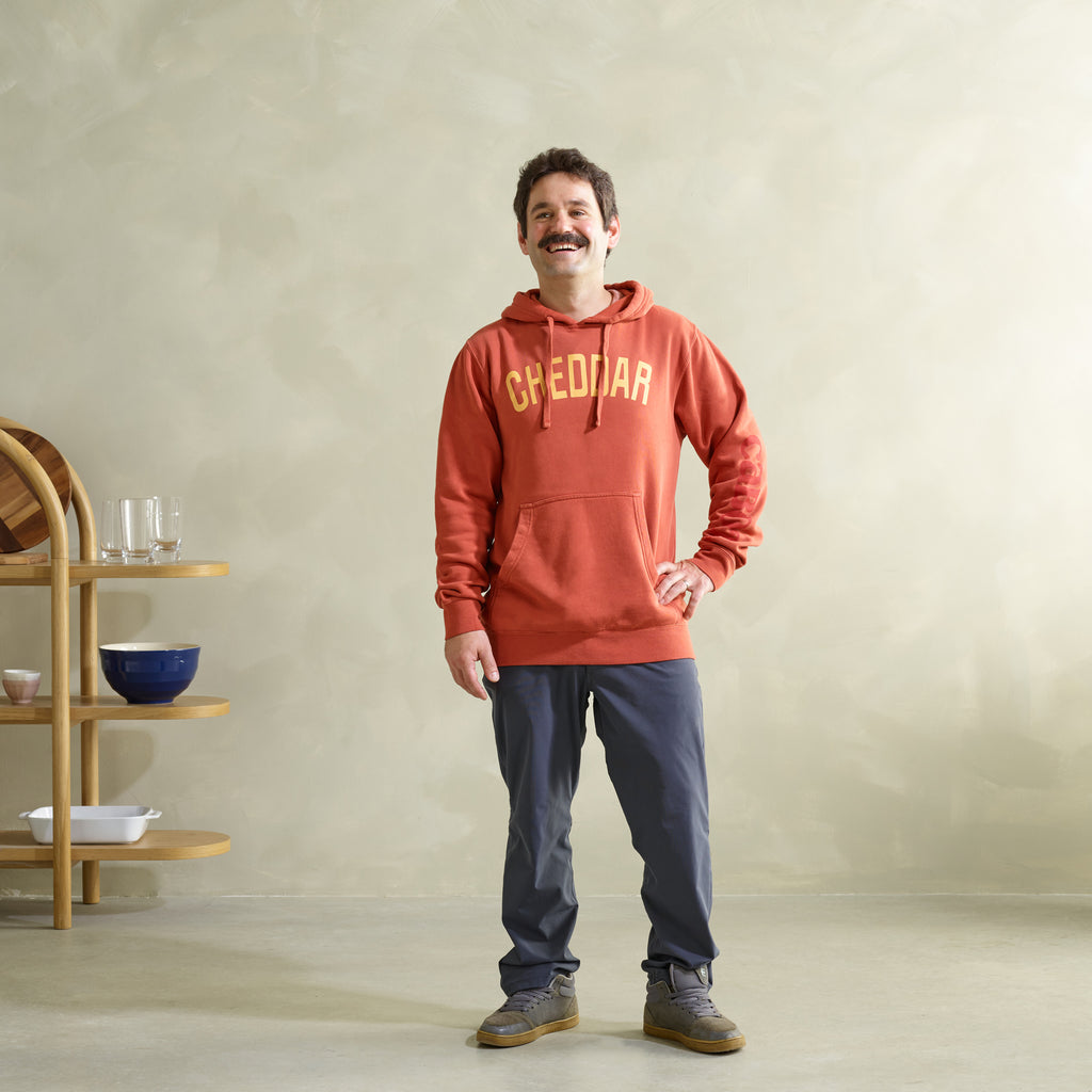 A mustachioed man smiles in a vibrant orange New Duds Cheddar Hoodie, showcasing its ultra-soft fabric and bold CHEDDAR print. Paired with blue pants and sneakers, he exudes vintage style against wooden shelves filled with bowls and glasses against a beige wall.