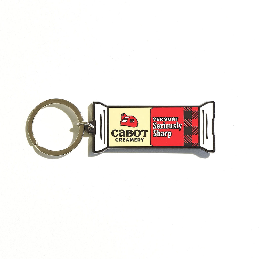 The Seriously Sharp Charm Keychain by WizardPins is shaped like a Cabot Creamery cheese package, featuring a Vermont Seriously Sharp label, red and black plaid design, and silver key ring—a fun accessory for cheese lovers.