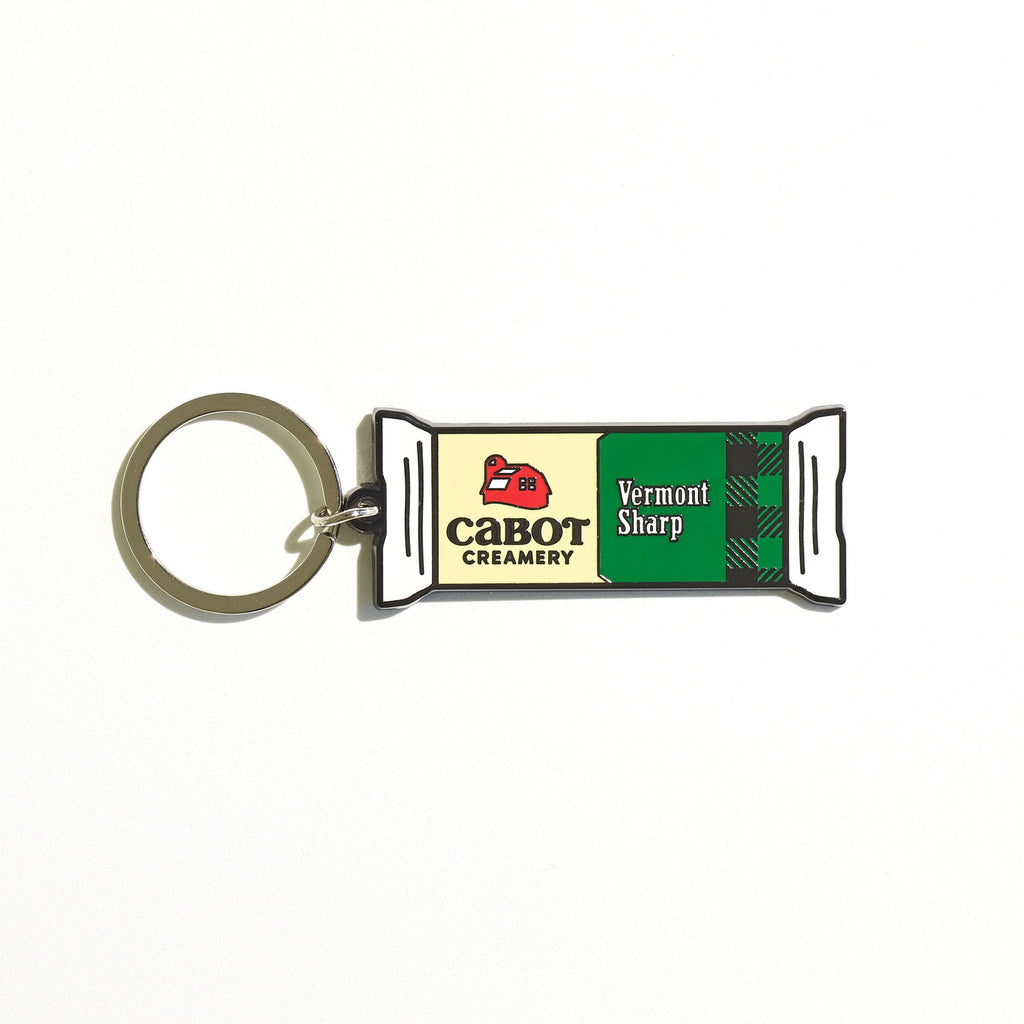 The Vermont Sharp Charm Keychain by WizardPins is shaped like a Cabot Creamery cheddar package, with the creamery logo and green plaid on a white background.