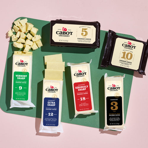 A Sharpness Tasting Cheese Sampler from Cabot Creamery is elegantly displayed on a green cutting board, featuring Vermont Sharp (9 months), Extra Sharp Cheddar (12 months), Seriously Sharp (18 months), Aged Creamery White (5 years), and Premium Cheddar (10 years) with cheese cubes adorning the board.