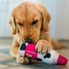 A golden retriever lies on a rug, intently chewing on the Cabot x Play Whipped Cream Crinkler Dog Toy by Play Pet Brands, a plush toy shaped like a milk bottle with a pink and black cow pattern.