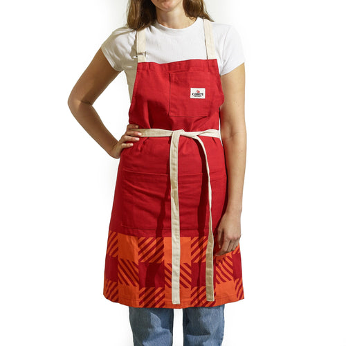 A person models the Gallant International Inc. Plaid Apron, featuring orange geometric patterns, a front pocket, and contrast straps, over a white t-shirt and blue jeans against a white background.