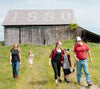 A family of five walks in a grassy area in front of an old wooden barn with 1880 on the roof. Two adults and three children, all casually dressed, are smiling and talking, enjoying the sunny day.