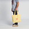 A person in a light blue shirt and dark pants holds the yellow Life is Butter Tote from New Duds. The plain, neutral background emphasizes the accessory’s cheerful vibe and Vermont-inspired style.