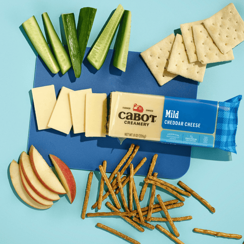 Sliced cucumbers, apple slices, Cabot Creamery Mild Cheddar Cheese, rectangular gluten-free crackers, and pretzel sticks are arranged on a blue background.