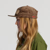 A person with long, light brown hair wears the New Duds Cheddar Corduroy Hat in Walnut and a green shirt, facing left against a plain light background.