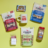 The Cozy Care Package from Cabot Creamery features an inviting gift box filled with assorted Cabot Creamery cheeses, a mug, chocolate chip cookies, cocoa mix, and maple candy—sweet and savory treats displayed on a yellow background.