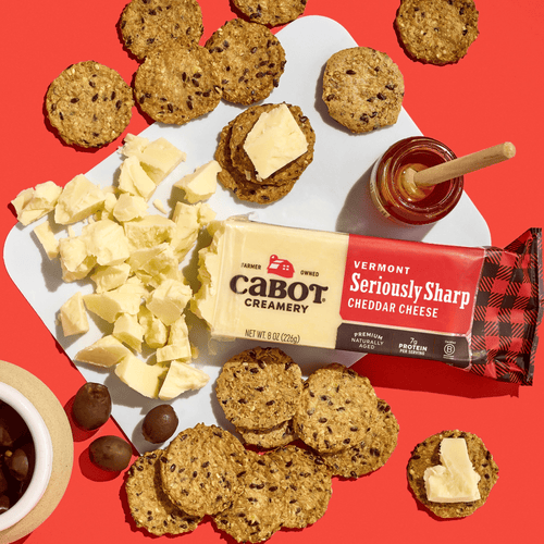 A block of Cabot Creamery Seriously Sharp Cheddar Cheese, accompanied by crackers, cheese chunks, a honey dipper with honey, olives, and a small bowl, is beautifully arranged on a white serving board against a vibrant red background.
