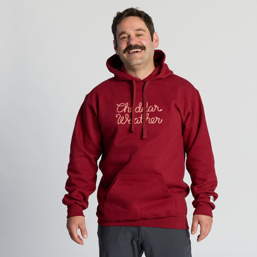 A man with a mustache smiles, wearing the New Duds Cheddar Weather Chainstitched Hoodie in maroon and dark pants, standing against a plain light background.