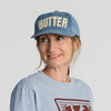 A woman wearing a New Duds Butter Corduroy Hat in blue, styled as an adjustable snapback with yellow lettering, and a matching blue shirt smiles and looks to the side against a plain background.