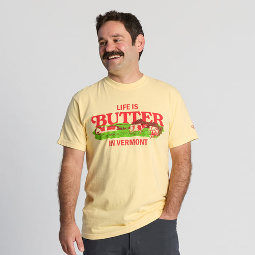 A man with a mustache is smiling and standing against a plain background, wearing the New Duds “Life is Butter in Vermont” T-shirt—a light yellow shirt featuring images of cows and a barn. He has one hand in his pocket.