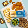 A cheese board featuring cubed Cabot Creamery Colby Jack Cheese, dried apricots, a bowl of green olives, and seeded crackers is arranged on a white plate against a light blue background. The award-winning Cabot Creamery Colby Jack Cheese brings extra delight to this elegant presentation.