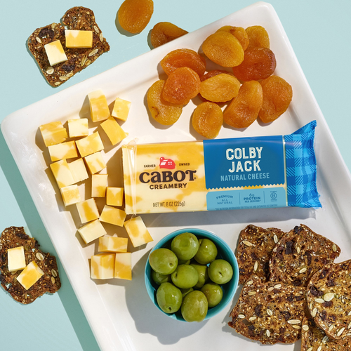 A cheese board featuring cubed Cabot Creamery Colby Jack Cheese, dried apricots, a bowl of green olives, and seeded crackers is arranged on a white plate against a light blue background. The award-winning Cabot Creamery Colby Jack Cheese brings extra delight to this elegant presentation.