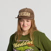A person wearing a walnut Cheddar Corduroy Hat from New Duds and a green CHEESE FROM VERMONT shirt smiles slightly against a plain background.