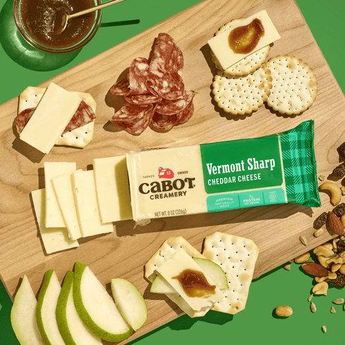 A wooden board featuring Cabot Creamery Vermont Sharp Cheddar Cheese, cheese slices, crackers, salami, pear slices, nuts, and fig jam is arranged neatly on a green background.