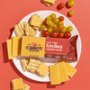 A white plate with sliced Cabot Creamery New York Extra Sharp Yellow Cheddar Cheese, square crackers, and red and yellow grape tomatoes rests on a red surface. A package of Cabot Creamery New York Extra Sharp Yellow Cheddar Cheese is also on the plate.