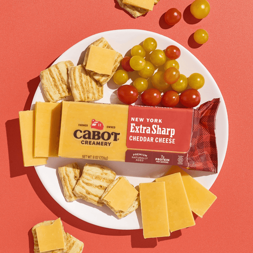 A white plate with sliced Cabot Creamery New York Extra Sharp Yellow Cheddar Cheese, square crackers, and red and yellow grape tomatoes rests on a red surface. A package of Cabot Creamery New York Extra Sharp Yellow Cheddar Cheese is also on the plate.