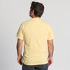 A man stands facing away from the camera, wearing the New Duds Life is Butter in Vermont T-shirt in light yellow and dark pants against a plain light background.