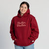A person smiles at the camera with their hands in the front pocket, wearing the New Duds Cheddar Weather Chainstitched Hoodie—a maroon sweatshirt featuring “Cheddar Weather” in script—while standing against a plain light background.