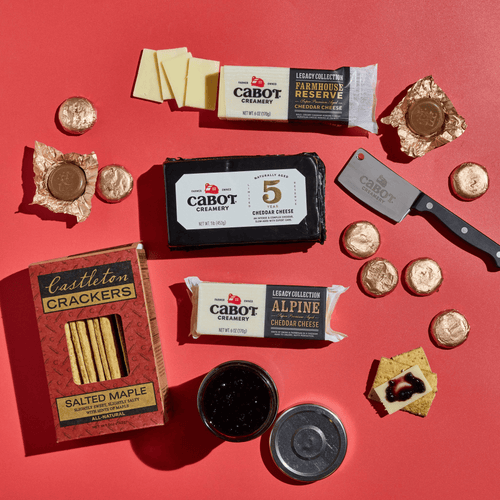 A Salty & Sweet Gift Pack featuring three blocks of Vermont cheeses, a box of salted maple crackers, a jar of fruit spread, a cheese knife, and several foil-wrapped chocolate candies arranged on a red surface.