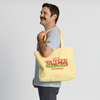 A smiling man with a mustache stands sideways, holding a pale-yellow Life is Butter Tote from New Duds on his shoulder. The tote features an illustration of cows and a farm, along with the phrase “Life Is Butter in Vermont.”.