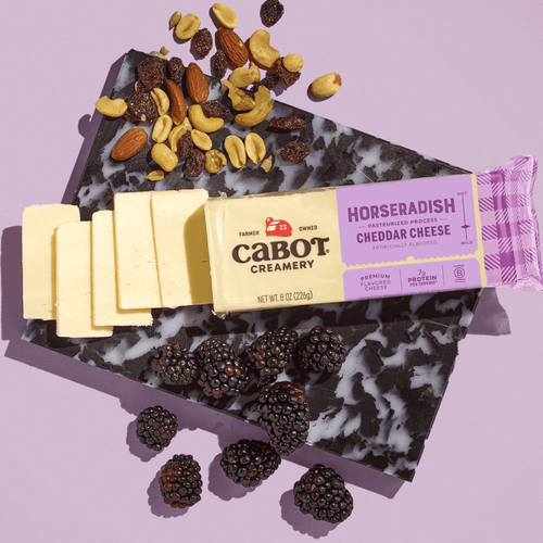 Cabot Creamery Horseradish Cheddar Cheese, a rich and lactose free cheese, is sliced and served with mixed nuts and blackberries, all artfully displayed on a marbled cutting board set against a light purple background.