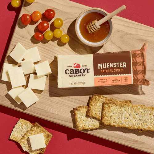 A wooden board with sliced creamy Muenster Cheese from Cabot Creamery, cherry tomatoes, whole grain crackers, and a dish of honey with a dipper, all arranged on a red background.