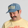 A smiling man with a mustache wears the Butter Corduroy Hat in blue by New Duds, which features cream “BUTTER” lettering, along with a yellow T-shirt with red text.