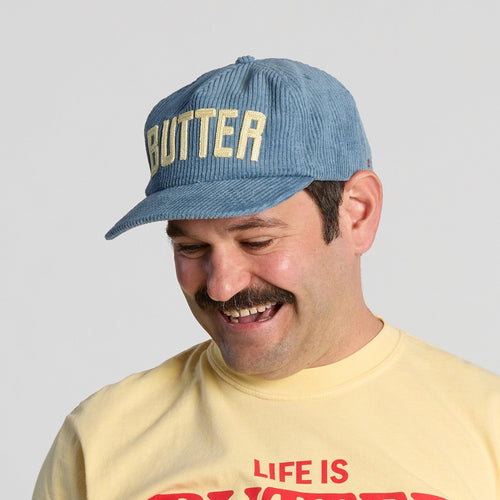 A smiling man with a mustache wears the Butter Corduroy Hat in blue by New Duds, which features cream “BUTTER” lettering, along with a yellow T-shirt with red text.