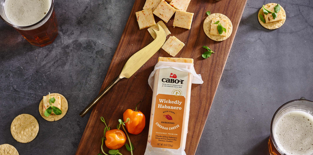 a bar of habanero cheddar on a cutting board with slices of cheese, crackers, habaneros, and micro greens, with two beverages.