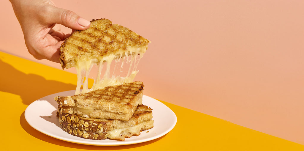 Pink and yellow background with a hand pulling up a half of a cheesy grilled cheese sandwich.