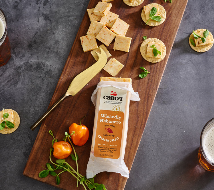 a bar of habanero cheddar on a cutting board with slices of cheese, crackers, habaneros, and micro greens, with two beverages.