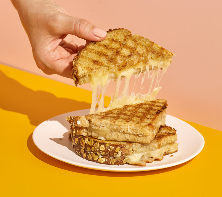 Pink and yellow background with a hand pulling up a half of a cheesy grilled cheese sandwich.