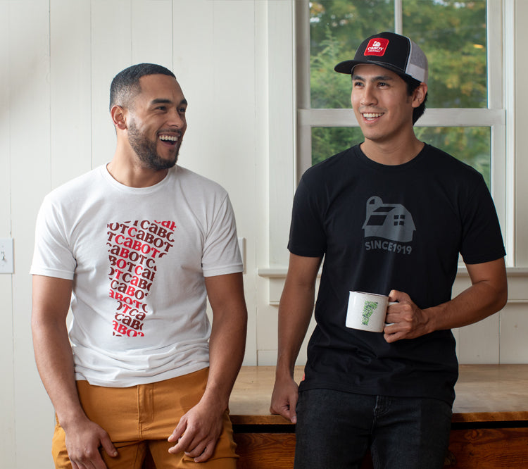 two men are standing in a kitchen, one is wearing a white cabot shirt, and the other is wearing a black cabot shirt and a cabot trucker hat, and holding a white cabot mug.