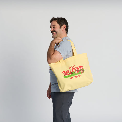 A smiling man with a mustache holds a Life is Butter Tote by New Duds, its bold red text and green accents celebrating Vermont’s dairy heritage and the Cabot cooperative, as he stands against a plain light background.