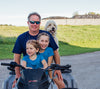 A man with sunglasses is sitting on an ATV with two smiling young girls in matching blue shirts in front of him. A fluffy dog is sitting behind them, poking its head over the mans shoulder. They are outdoors on a sunny day with grass in the background.