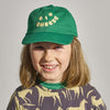 Smiling child with shoulder-length hair wears the New Duds Big Cheese Youth Hat (green with “BIG CHEESE” in yellow) and a horse-patterned shirt.