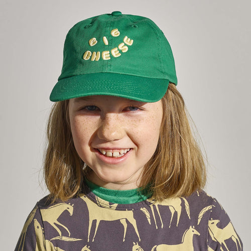 Smiling child with shoulder-length hair wears the New Duds Big Cheese Youth Hat (green with “BIG CHEESE” in yellow) and a horse-patterned shirt.