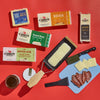 The Cabot Creamery Raclette and Cheese Gift Set features award-winning cheeses, a slicer with melted cheese, salami slices, a spread jar, and a blue cutting board with cheese and knife—all on a bold red background.