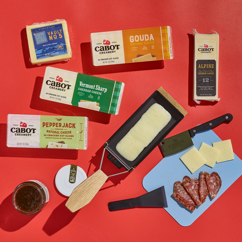 The Cabot Creamery Raclette and Cheese Gift Set features award-winning cheeses, a slicer with melted cheese, salami slices, a spread jar, and a blue cutting board with cheese and knife—all on a bold red background.