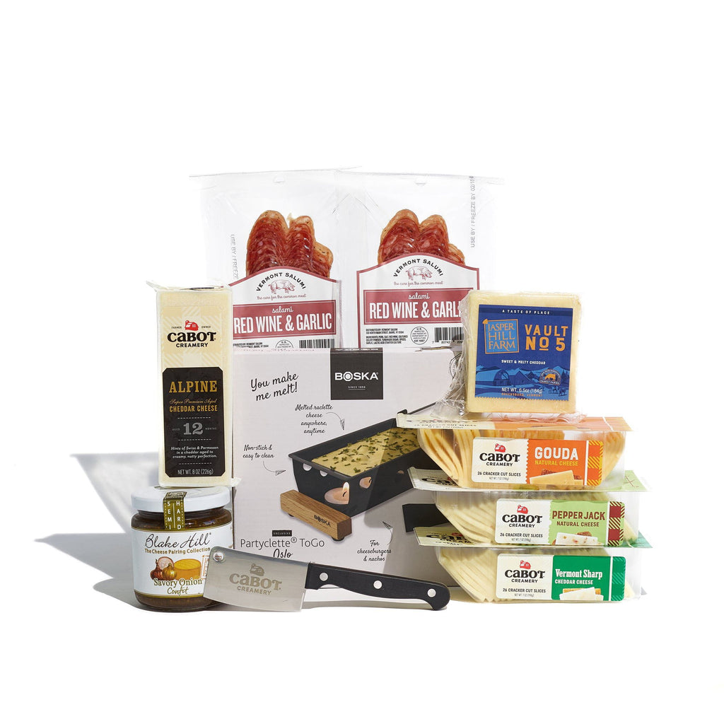 The Raclette and Cheese Gift Set by Cabot Creamery includes award-winning cheeses, red wine and garlic sausages, a cheese knife, a mini serving board, and Blake Hill preserve—all beautifully arranged against a white background.