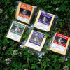 Five packages of Sweet Rowen Farmstead cheese arranged on clover grass. Varieties include Horseradish Chive Cheddar, Herb Cheddar, Mild Cheddar, Aged Cheddar, and Hot Cheddar. Each package features a unique label.