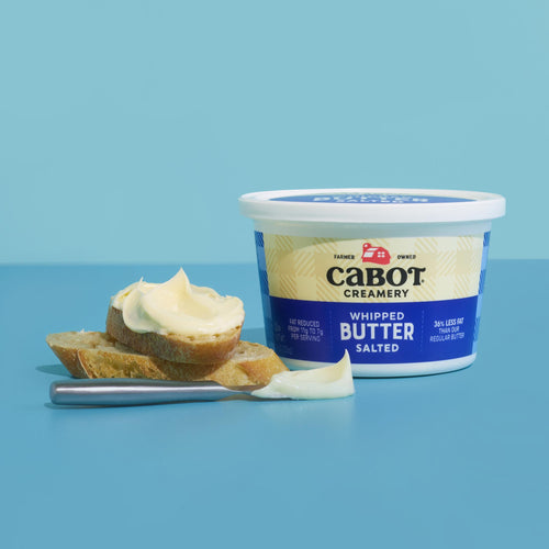 A container of Cabot Creamery Whipped Salted butter is placed beside two buttered bread slices and a knife, all on a blue surface with a blue background.