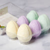 A clear plastic carton holds six egg-shaped bath bombs. The bath bombs are in pastel colors: two yellow, two purple, and two light blue-green. They are arranged in two rows and sit on a marble countertop.