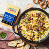 A skillet with cheesy dip topped with bacon, green onions, and mustard is surrounded by sliced bread, pretzels, and herbs. A package of Cabot Creamery Extra Sharp Cheddar Cheese sits beside it.