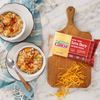 Two bowls of macaroni and cheese with herbs and paprika are on a cloth next to a wooden board displaying a block of Cabot Creamerys New York Extra Sharp Yellow Cheddar Cheese and shredded cheese on marble.