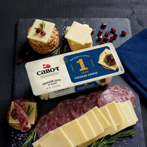 Slices of Cabot Creamerys 1 Year Cheddar Cheese and salami are presented on a tray. Nearby, crackers with cheddar cuts, rosemary, and cranberries are arranged. The scene is decorated with a blue napkin and sprigs of rosemary.